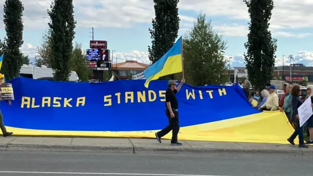 People hold a large sign on a Ukrainian flag reading: Alaska stands with.