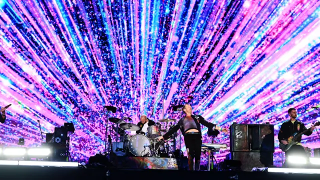 A band playing drums and guitars stand on a stage. A large screen covered in blue, white and pink lights is in the background.