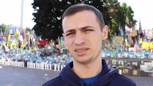 Man with dark hair wearing blue sweatshirt with a display of Ukrainian flags behind him