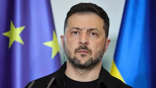 Close up of Volodymyr Zelensky wearing a black top as he stands in between a EU flag to his right and a Ukrainian flag to his left