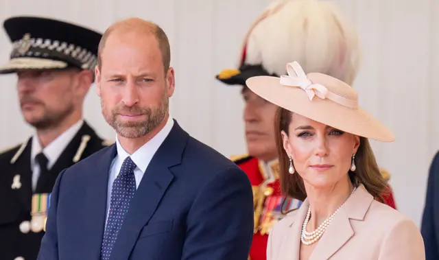 William and Kate stand next to each other at an event. He wears a blue suit and spotted tie, she wears a faded pink blazer and hat.