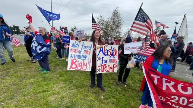 A group of Trump supporters holds up pro-Trump signs and flags. A woman (C) with glasses stands holding two signs, one in her right hand reading "Not tired of winning bigly", another in her left hand reading "Alaska for Trump"