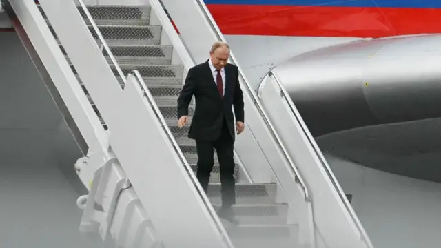 Vladimir Putin in a black suit walks down the steps of the stairs of his presidential plane after landing in Alaska