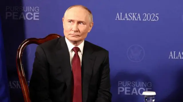 Putin wearing a red tie, white shirt and black suit, sat on a brown wooden chair, in front of a backdrop with the words Alaska 2025 and Pursuing Peace