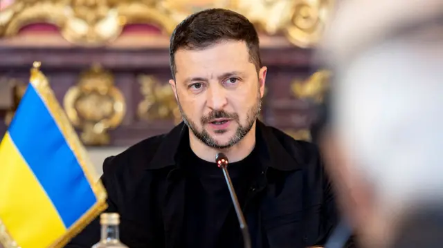 Volodymyr Zelensky speaks into microphone, he's sitting next to a Ukraine flag
