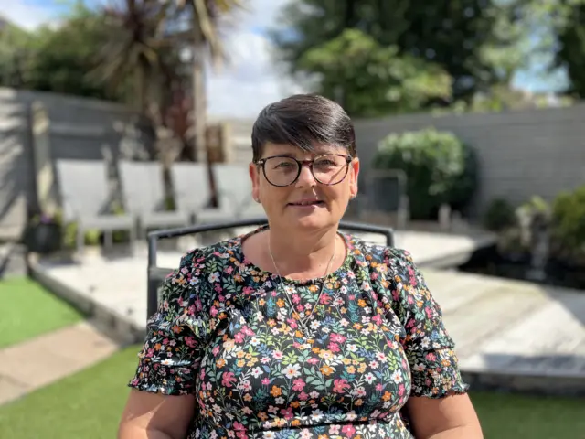 Cheryl Cox sitting in chair in her sunny garden, she has short dark hair and tortoiseshell glasses and is wearing a floral dress. Her garden behind her has lawn and a deck area with chairs and shrubs in pots