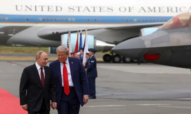 Trump and Putin walking on a red carpet, both wearing dark suits with red ties. Behind them in a runway and planes with soldiers stood holding flags. The pair are walking close to each other and talking.