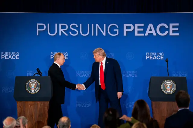 Vladimir Putin, Russia's president, left, and US President Donald Trump shake hands during a joint news conference in Alaska. In front of them are two presidential lecterns with the US government seal. Behind them is a big blue background with the words: "Pursuing Peace" written in white.