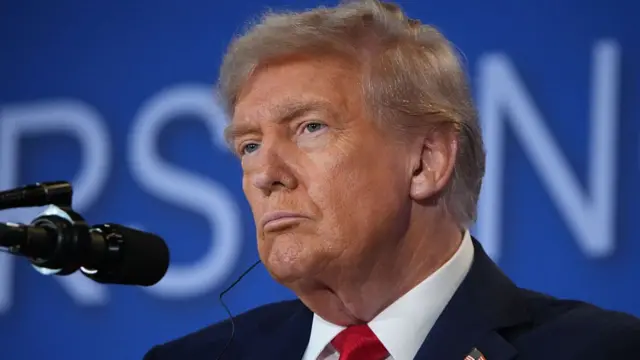 President Trump in a dark suit with white shirt and red tie, standing behind a microphone, blue background behind him.