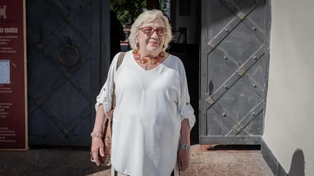 Valentyna Sikora standing in a long white t-shirt with a large red and orange necklace and glasses. She stands in front of a doorway holding a bag with arms by her side, she is smiling and looking to the right.