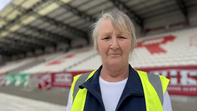Anne is wearing a hi-vis vest over a blue fleece gilet and white t-shirt, she's standing in front of the stands at Craven Park - the seats spell E R in red and white