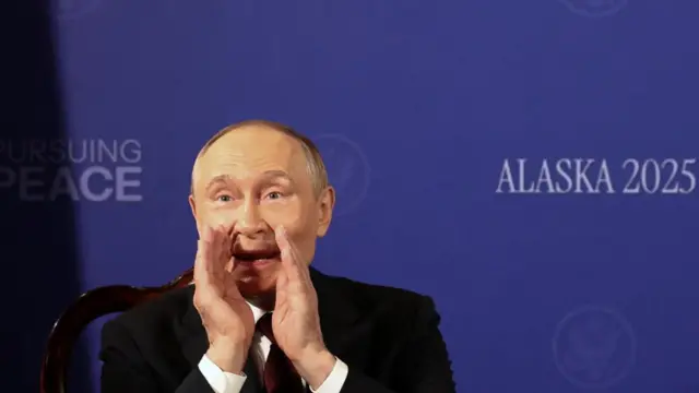 Putin, sitting in front of the blue Alaska 2025 background, cups his hands to his mouth as he shouts something to someone off camera