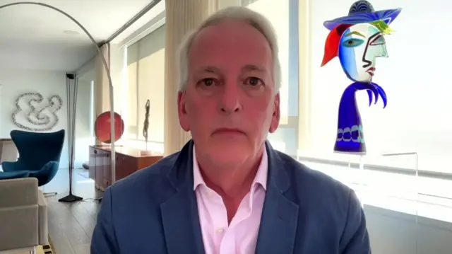 Ivo Daalder, elderly man with white hair sitting down indoors. He's wearing a blue-grey blazer with a pink shirt , behind him several pieces of art are visible decorating the room