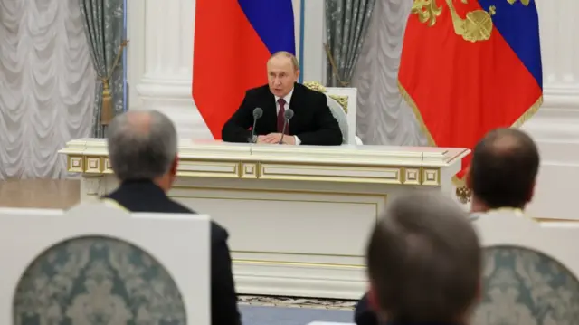 Vladimir Putin (C) sits down at a white wooden desk with gold decorations as he sits on a matching chair in front of Russian officials at the Kremlin. Behind him, affixed to the wall, is a Russian flag
