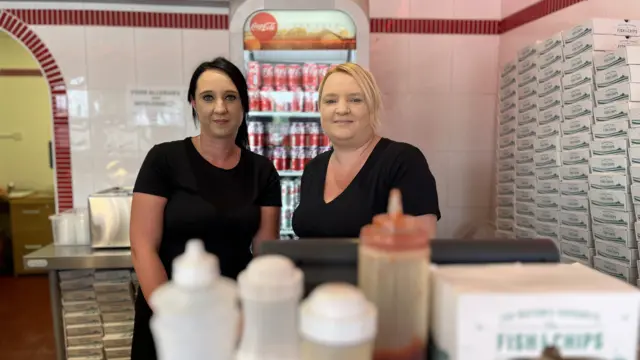 Katy and Sam stand behind the till in their fish and chip shop, stacks of cardboard trays are lining the wall to their right. behind them is a an illuminted fridge filled with drinks cans. In front of the till and out of focus are traditional plastic salt and vinegar shakers and a tomato sauce bottle, The sisters both have black t-shirts on and have their hair in ponytails, Katy on the left is dark haired and Sam is blonde.