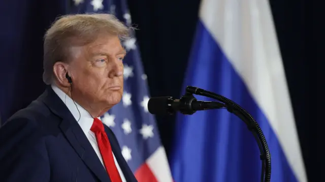 Mid-shot of Donald Trump in a dark suit (L) standing on stage in front of black mic. In the blurred background behind him are a US and Russian flag