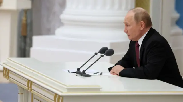 Wide shot from the left side of Vladimir Putin in a black suit sitting down at a white wooden desk decorated in gold speaking into two mics