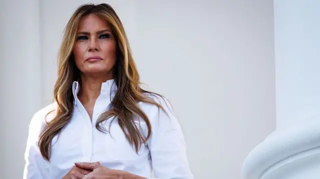 Melania Trump in a white button-up shirt looking into the distance. Her hands are clasped together.
