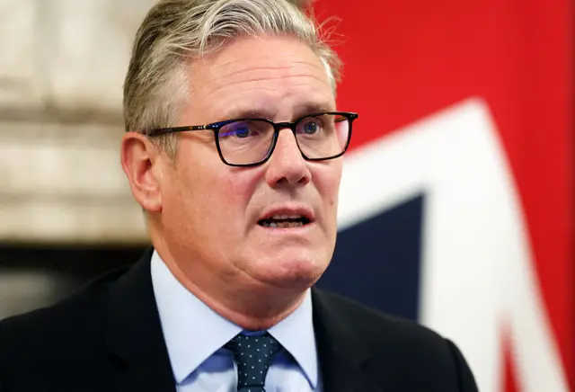 UK Prime Minister Keir Starmer in suit, tie and glasses. UK flag behind him.