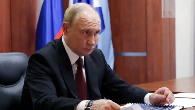 Vladimir Putin in a black suit, white shirt and burgundy tie sitting on a leather chair at a wooden desk, a Russian flag on a pole behind him next to a wood-panelled wall