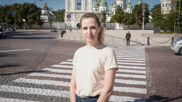 Maria Drachova in a white t-shirt and jeans stood on the pavement, behind her is a road with traffic lights and pedestrian crossing. A man stand on the other side of the road in the distance.