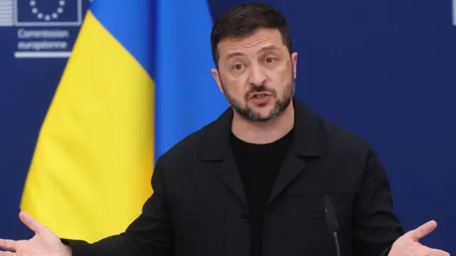 Close up of Volodymyr Zelensky in black blazer and black shirt, gesturing with his hands, a Ukrainian flag on a pole behind him