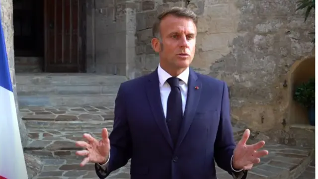 Mid-shot of Emmanuel Macron in dark blue suit, black tie and white shirt in front of the steps of an old building, a French flag to his right
