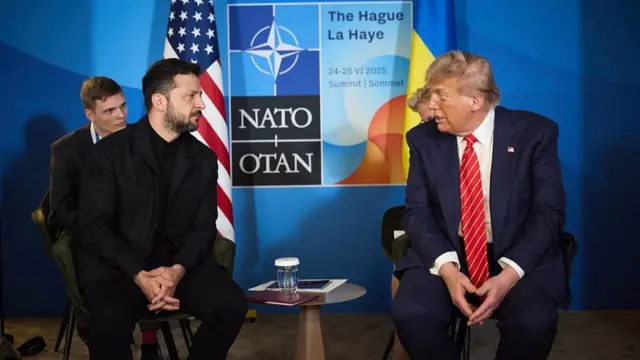 Volodymyr Zelensky (L) clad in black sits on a grey chair with Donald Trump (R) sitting on a matching chair to his left. They both have their hands crossed in front of them as they chat