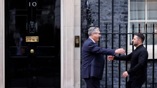 Keir Starmer and Volodymyr Zelensky outside 10 Downing Street. They are walking towards each other with arms out-stretched, about to hug. Both smiling, railings and a black door behind.