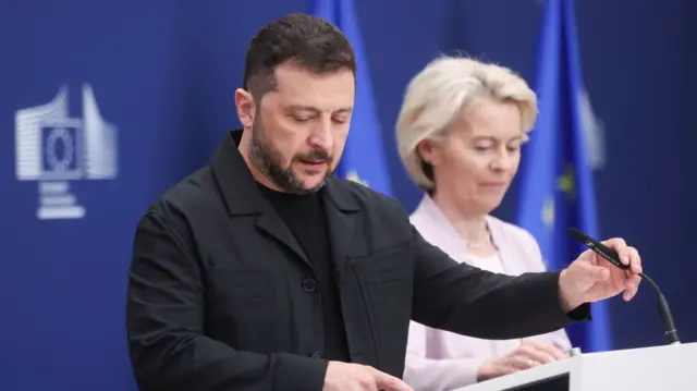 Zelensky, wearing a black t-shirt and black jacket, speaks into a microphone while a finger on his right hand guides him through his speech written on the paper in front of him