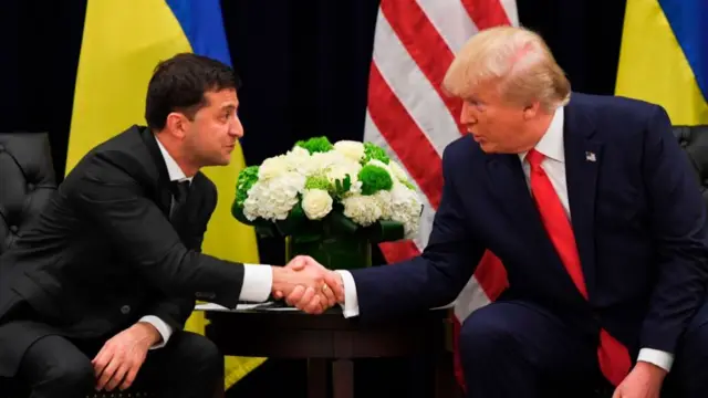 Volodymyr Zelensky (L) in a black suit shakes hands with Donald Trump (R) also in a suit while they're both sitting down. A low table with a flower arrangements stands between their chairs, two Ukrainian flags and a US one behind them