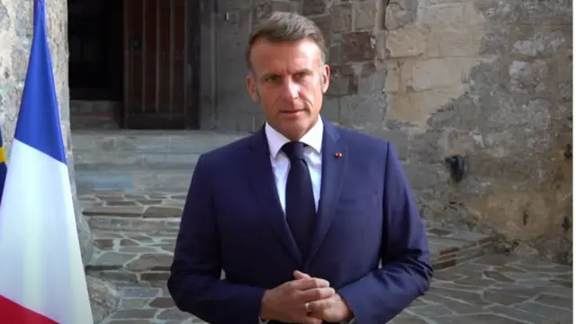 Close up of Emmanuel Macron in dark blue suit, white shirt and black tie with his hands crossed in front of him as he stands in front of an old building, a French flag to his left