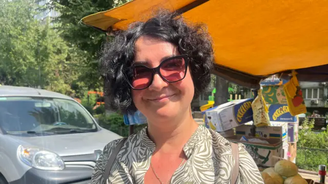 Iryna Levchuk smiles at the camera in the sun. She's wearing large black sunglasses with pink-tinted lenses and a grey and cream shirt. A Grey car is parked to her right and there's a fruit and veg stall behind her