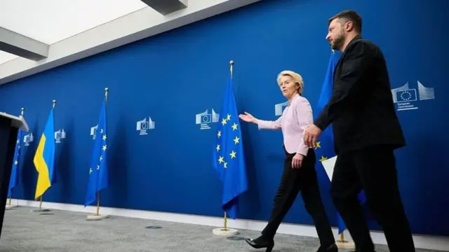 Ursula von der Leyen in a lilac blazer and black trousers walks slightly ahead of Volodymyr Zelensky before a joint news conference. They're walking past a blue wall covered with the European Commission logo and lined with EU and Ukrainian flags
