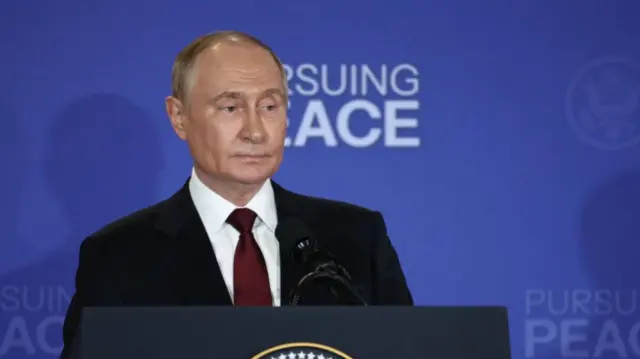 Vladimir Putin mid-shot, he's in a black suit, burgundy tie and white shirt. Behind him a blue wall with writings reading Pursuing peace in white