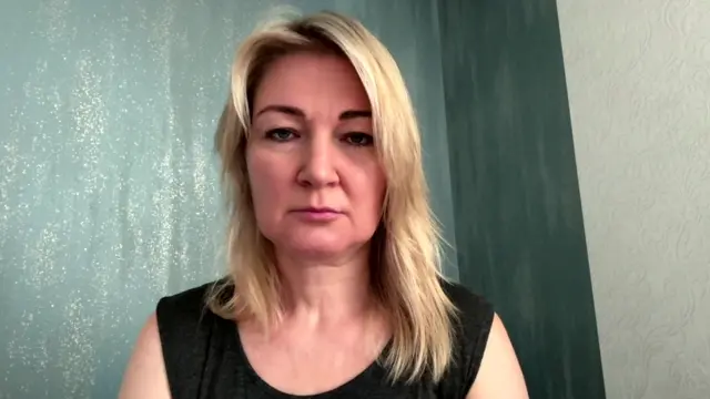 Mariya Ionova looks directly at the camera. she has long blonde hair which is slightly longer over her left shoulder. She is wearing a black top and has a whie and green backdrop behind her