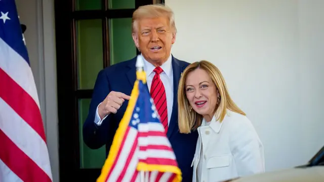 Donald Trump (L) and Giorgia Meloni (R) stand close to each other outside the White House as he welcomes her for a visit. He's in a dark blue suit with a striped red tie, while she's in a white jacket and matching shirt, two US flags visible to their sides