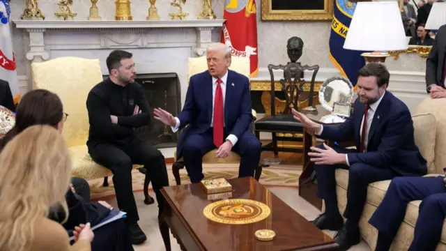 Wide shot of the Oval Office. To the left, Volodymyr Zelensky in black clothing with his arms crossed in front of him as he sits on a yellow brocade chair to the right of Donald Trump. Trump (C) sits on a matching chair in a dark blue suit, red tie and white shirt as he gestures towards Zelensky with his right hand. JD Vance, sitting on a yellow couch, looks to his left as he gestures with both hands towards the duo