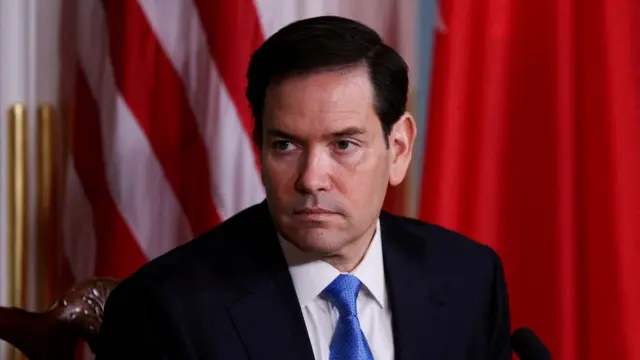 Close up of US Secretary of State Marco Rubio sitting down on a wooden chair wearing a dark blue suit, white shirt and light blue tie. Behind him a US flag and red curtain