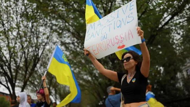 A woman holds up a sign reading "massive sanctions on terrorussia", with letters in Russia dripping with blood. People hold Ukrainian flags behind her
