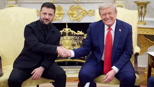 Zelensky, in a black suit with black shirt, and Trump in a blue suit with a red tie, smile and shake hands sat on yellow chairs in the Oval Office