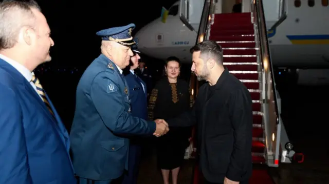 Zelensky in a black jacket shakes hands with officials while it is still dark