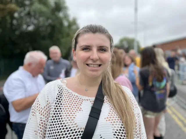 A head and shoulders shot of a woman - Lydia Gaftarnik - smiling. She has long blonde hair tied in a ponytail. She is wearing a black vest top with a white crochet jumper over the top. She is also wearing a bag with a black strap across her chest and a silver necklace. Crowds can be seen blurred in the background.