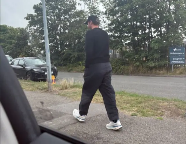 A man wearing a black baseball cap, black jumper, grey trousers and white trainers walks away from the camera along a pavement