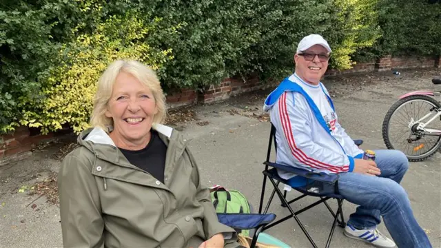 A man and a woman sat on blue deckchairs on a path, smiling at the camera. Bushes can be seen behind them. The woman has short blonde hair and is wearing a black t-shirt and a green waterproof coat. The man is wearing blue jeans, white and blue trainers, a grey jacket with three red stripes down the arm, a white t-shirt, sunglasses and a white cap.