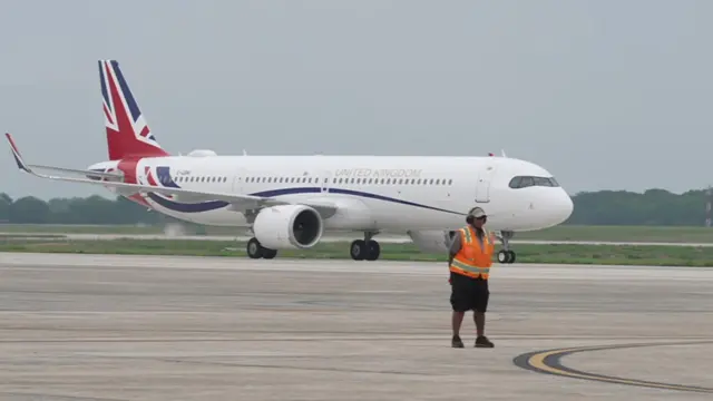 Plane with Union Jack flag on its tails taxis on tarmac, an airport employee in an orange hi-vi jacket waits on the ground