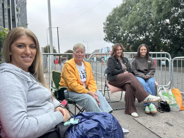 Four ladies look at the camera as they sit in deck chairs surrounded by metal barriers at the side of a road. One woman has long dark blonde hair and a grey hoodie, another has short blonde hair with a yellow hoodie and blue jeans. Another lady has a black jacket on with brown trousers and another with a grey hoodie and blue jeans.