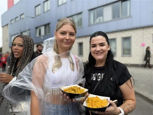 Head and shoulders shot of two women -  Sarah Andrew and Sarah Murray - stood in a street, smiling at the camera holding chips in a cardboard tray. The woman on the left is wearing a transparent rain poncho, blue glittery trousers and a white crop top. She has long blonde hair. The woman on the right has long black hair and is wearing a black t-shirt.