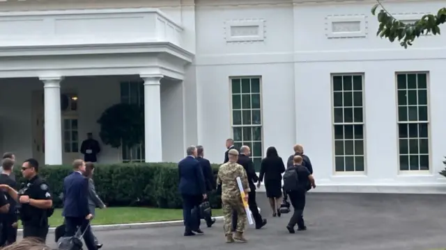 A man in army attire walking into the White House carrying a long white tube
