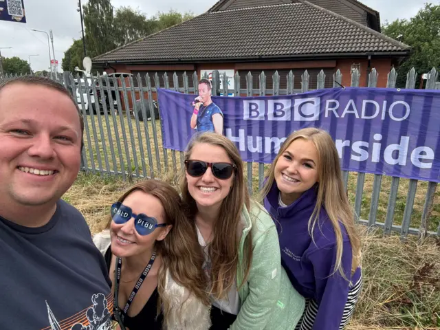 Three people, one man and three women standing on grass outside. There is a purple banner reading "BBC RADIO HUMBERSIDE" hung on metal railings. A cardboard cut out of Chris Martin is also behind them.